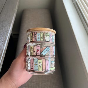 May include: A clear glass jar with a wooden lid and a colorful design of books on a bookshelf. The books have titles like "Bloom", "Trees", "Flower", "Spring", "Book Lover", "Spicy", "Read", and "Spring!! Vol. 2".