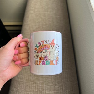 Puede incluir: Taza de cer&aacute;mica blanca con interior y asa rosas. La taza presenta un colorido dise&ntilde;o de Halloween con un fantasma, un arco&iacute;ris, un sombrero de bruja y las palabras "Stay Spooky".