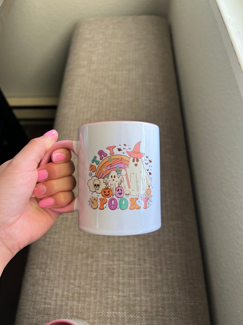Puede incluir: Taza de cer&aacute;mica blanca con interior y asa rosas. La taza presenta un colorido dise&ntilde;o de Halloween con un fantasma, un arco&iacute;ris, un sombrero de bruja y las palabras "Stay Spooky".