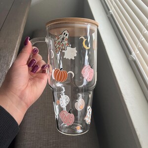 May include: A clear glass mug with a handle and a bamboo lid, decorated with Halloween-themed stickers. The stickers include pumpkins, ghosts, and a crescent moon. The mug is held by a person.