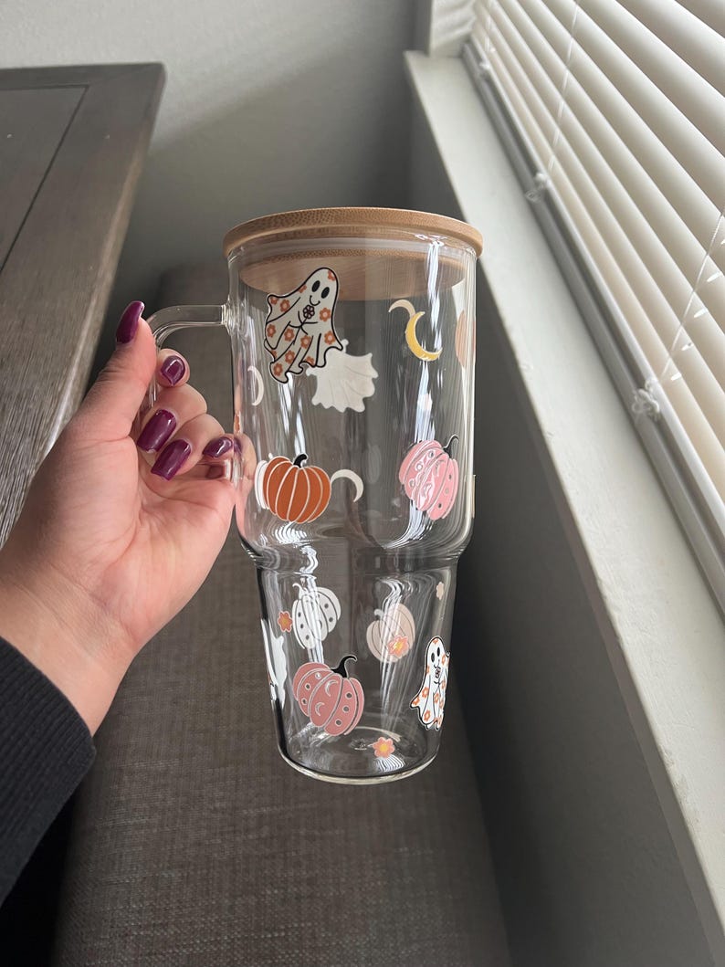 May include: A clear glass mug with a handle and a bamboo lid, decorated with Halloween-themed stickers. The stickers include pumpkins, ghosts, and a crescent moon. The mug is held by a person.