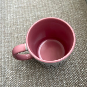 Puede incluir: Una taza de cer&aacute;mica rosa con un asa a juego. El interior de la taza es de un tono rosa m&aacute;s oscuro. El exterior de la taza es gris con letras blancas. La taza est&aacute; sobre una superficie texturizada.