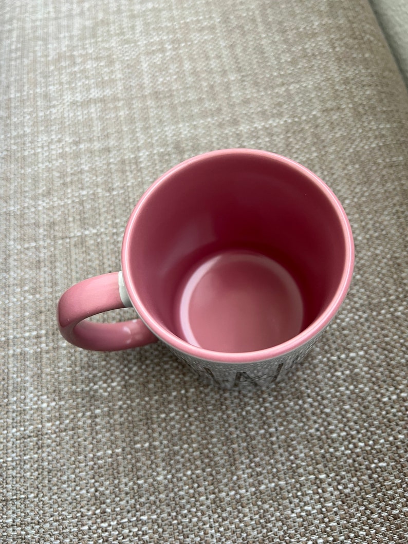 Puede incluir: Una taza de cer&aacute;mica rosa con un asa a juego. El interior de la taza es de un tono rosa m&aacute;s oscuro. El exterior de la taza es gris con letras blancas. La taza est&aacute; sobre una superficie texturizada.