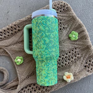 May include: A green and yellow bedazzled tumbler with a white lid and a mint green handle. The tumbler has a white straw. The tumbler is next to a brown knitted bag and three small crochet flowers.