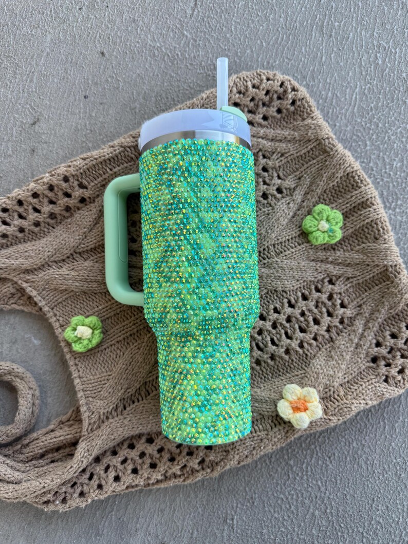 May include: A green and yellow bedazzled tumbler with a white lid and a mint green handle. The tumbler has a white straw. The tumbler is next to a brown knitted bag and three small crochet flowers.