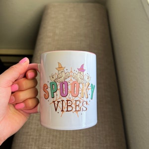 Puede incluir: Taza de cer&aacute;mica blanca con interior y asa rosas, con el texto "SPOOKY VIBES" en letras coloridas. El dise&ntilde;o incluye fantasmas de dibujos animados con sombreros de bruja y una telara&ntilde;a. Una taza con tem&aacute;tica de Halloween.