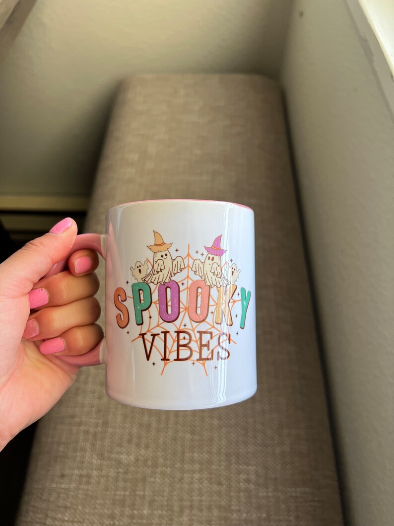 Puede incluir: Taza de cer&aacute;mica blanca con interior y asa rosas, con el texto "SPOOKY VIBES" en letras coloridas. El dise&ntilde;o incluye fantasmas de dibujos animados con sombreros de bruja y una telara&ntilde;a. Una taza con tem&aacute;tica de Halloween.