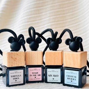 May include: Four car air fresheners with black Mickey Mouse-shaped tops, wooden blocks, and black square bottles. Each bottle has a different label with text: "Tortuga Tides", "Magic in the Air", "Sivako", and "Chateau Riviera".