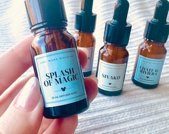 Könnte beinhalten: Vier braune Glastropfflaschen mit schwarzen Verschlüssen, jede mit einem hellblauen rechteckigen Etikett. Die vordere Flasche ist mit 'LIFE MADE MAGICAL', 'SPLASH OF MAGIC' und '10 mL DIFFUSER REFILL' sowie einem kleinen Maus-Symbol beschriftet.