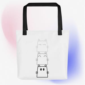 May include: White tote bag with black handles and a design of three stacked cats. The top cat is black with white eyes, the middle cat is gray with white eyes, and the bottom cat is gray with black eyes.