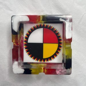 Puede incluir: Cenicero cuadrado con un diseño colorido. El centro presenta un cuadrado blanco con un emblema circular dividido en cuatro cuadrantes: blanco, rojo, negro y amarillo. El borde exterior es una mezcla de resina roja, amarilla, negra y blanca.