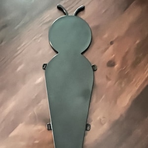 May include: A dark gray metal garden stake in the shape of a stylized insect. The stake has a rounded head with two antennae and a long, tapered body. Small loops are attached to the body for securing the stake.