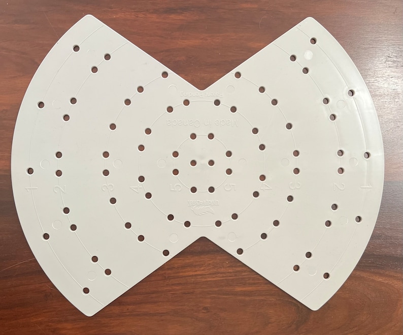 May include: A light grey plastic craft tool with numerous evenly spaced holes and curved edges. The tool is shaped like two semi-circles joined at a point. The text "Made in Canada" is visible.