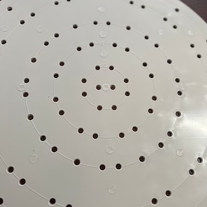 May include: A white plastic craft circle with concentric rings of evenly spaced holes. Numbers 2 through 5 are printed along the rings. The circular design is set against a dark wood surface.