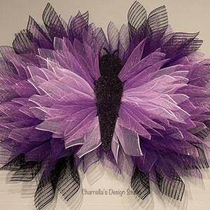 May include: A butterfly-shaped wreath crafted from purple, black, and white mesh. The wings display gradients of purple, fading to black at the edges. The central body is black and glittery. Designed for wall hanging, it's a decorative piece.