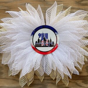 May include: A white and gold mesh wreath with a central circular emblem. The emblem features the text "9/11 Never Forget" with a silhouette of the Twin Towers, the Statue of Liberty, and the American flag. The wreath is set against a wooden background.