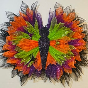 May include: A decorative butterfly wreath with a glittery black body and wings made of layered orange, green, purple, and black mesh. Small spider decorations are attached to the wings. The wreath is designed for Halloween.