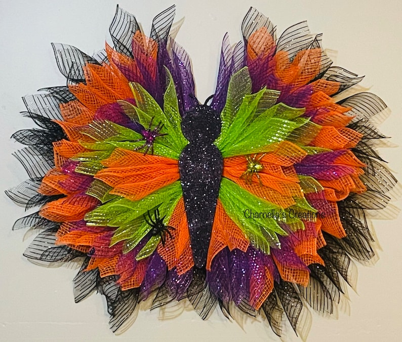 May include: A decorative butterfly wreath with a glittery black body and wings made of layered orange, green, purple, and black mesh. Small spider decorations are attached to the wings. The wreath is designed for Halloween.