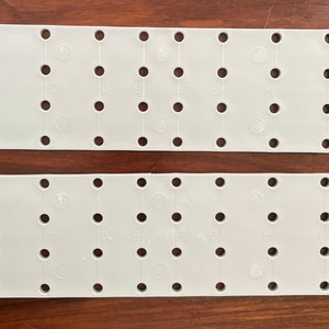 May include: Two gray plastic pegboards with rows of holes and letters labeled A through H on the top board and A through L on the bottom board.