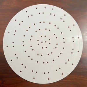 May include: A white, circular wreath-making form with concentric circles of evenly spaced holes. Numbers are printed on the form to indicate the size of the wreath. The form is made of a durable plastic material.