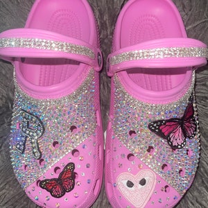 Pode incluir: Crocs rosa com uma tira de strass e vários enfeites, incluindo borboletas e um coração.