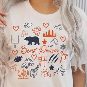 Puede incluir: Camiseta blanca con un dise&ntilde;o gr&aacute;fico que presenta un oso azul marino, un bal&oacute;n de f&uacute;tbol y otros iconos relacionados con el deporte. La frase "Bear Down" est&aacute; escrita en gui&oacute;n naranja, con acentos naranjas y azul marino adicionales.