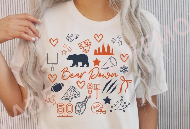 Puede incluir: Camiseta blanca con un dise&ntilde;o gr&aacute;fico que presenta un oso azul marino, un bal&oacute;n de f&uacute;tbol y otros iconos relacionados con el deporte. La frase "Bear Down" est&aacute; escrita en gui&oacute;n naranja, con acentos naranjas y azul marino adicionales.