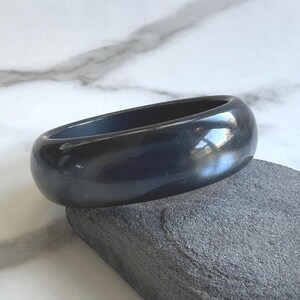 May include: A smooth, polished, solid black bangle bracelet with a rounded, thick profile. The surface of the bangle is highly reflective, showing subtle highlights and a deep, uniform dark color throughout the entire circular band structure.