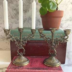 May include: A brass candelabra with an ornate, scrolled design and a fluted base. The candelabra features five candle holders, with three white taper candles inserted into the left and center sockets, while the right side remains empty.