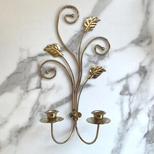 May include: A gold-toned metal wall sconce featuring two candle holders at the base. The structure includes curved wire stems that extend upward, decorated with three metallic leaves and two spiral accents, centered by a small flower detail.