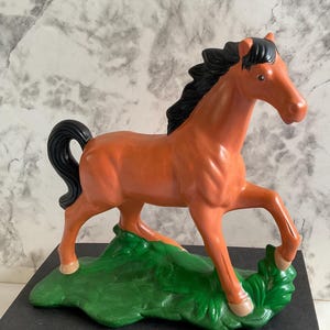 May include: A ceramic horse figurine in a trotting pose. The horse is a warm orange-brown color with a black mane and tail. It stands on a green base with a grassy texture. The figurine is placed on a book.