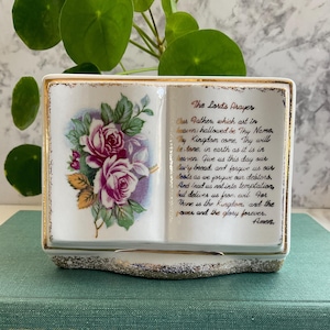 Puede incluir: Un joyero de cerámica blanca en forma de libro de oraciones con ribete dorado. El lado izquierdo presenta un diseño floral con rosas rosas y moradas. El lado derecho muestra el texto de "The Lord's Prayer".
