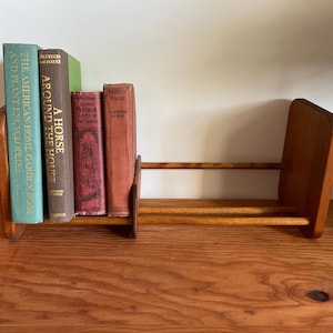 May include: A wooden book rack with two vertical end panels and two horizontal rods, holding four hardcover books titled The American Home Garden Book and Plant Encyclopedia, A Horse Around the House, Little Pictorial Lives of the Saints, and Saddle Bags.