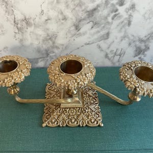 May include: A gold-toned metal candelabra featuring three circular candle holders with embossed floral detailing. The central base is square and also decorated with intricate floral patterns, connected to two side arms with a twisted metal design.