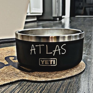 May include: A matte black stainless steel pet bowl with a silver metallic rim. The name ATLAS is engraved in a simple font above the YETI brand logo, which is enclosed in a rectangular frame on the front of the bowl.
