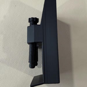 May include: A matte black C-clamp style mounting bracket featuring a vertical main body with a flat base and a top section equipped with a threaded adjustment screw and a circular knob for tightening against a surface to secure items in place.