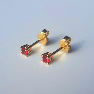 Puede incluir: Un par de pendientes de botón dorados, cada uno con una pequeña gema roja de corte cuadrado. Los pendientes tienen un diseño clásico de poste y cierre de mariposa.