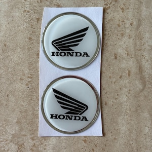 May include: Two circular Honda emblems featuring a black wing logo and the word HONDA in black text on a white background. Each emblem has a metallic silver border and is presented on a white rectangular backing sheet.
