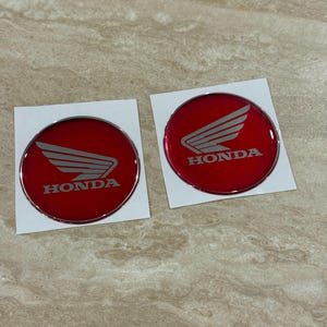 Può includere: Due emblemi circolari Honda con sfondo rosso lucido, un logo ad ala argentato e la parola HONDA stampata in argento. Ogni emblema è montato su un supporto adesivo bianco quadrato, caratterizzato da una finitura metallica e bordi netti.