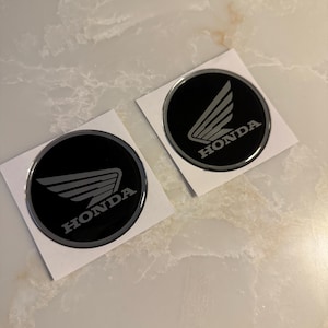 May include: Two black and silver Honda emblem stickers. Each sticker is round with a black background and the Honda logo in silver. The logo features a stylized wing design with the word "HONDA" below it in a silver, sans-serif font.