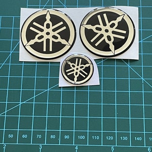 May include: Three black and cream-colored Yamaha logo stickers of varying sizes are displayed on a green cutting mat. The stickers feature a circular design with a stylized emblem. The stickers are on a white backing.