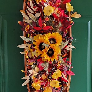 May include: A wooden oval-shaped basket filled with a fall floral arrangement. The arrangement features sunflowers, red and yellow flowers, and greenery. The basket is hanging on a green door.