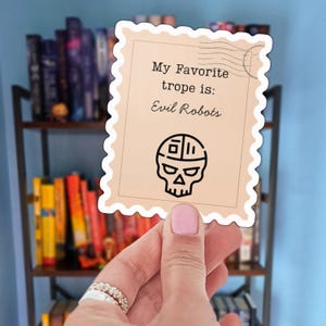May include: A sticker in the shape of a postage stamp with the text "My Favorite trope is: Evil Robots" and a skull-shaped robot design. The sticker is held in front of a bookshelf.