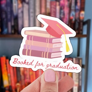Könnte beinhalten: Ein Aufkleber mit einem Stapel Bücher und einer rosa Abschlusskappe. Der Aufkleber hat den Text "Booked for graduation" in einer Schreibschrift. Die Bücher sind in Rosa- und Lilatönen gehalten.