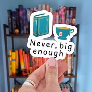 以下が含まれることがあります： ターコイズブルーの本とティーカップが描かれたステッカーで、「Never big enough」の文字が入っています。本棚の前でステッカーが持たれています。ティーカップにはハート型のタグが付いています。