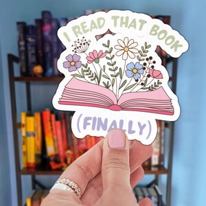 May include: A sticker with the text "I READ THAT BOOK (FINALLY)" in a whimsical font. The sticker features an open pink book with colorful flowers and foliage sprouting from it. The background shows a bookshelf.