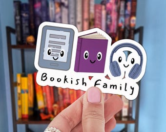 May include: A white sticker with the text "Bookish Family" and illustrations of an e-reader, a purple book, and headphones, all with smiling faces. The sticker is held in front of a bookshelf.