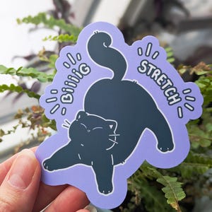 May include: A sticker featuring a black cat stretching on a light purple background. The sticker has the words "Biiiiig Stretch" in white, stylized text. The cat is in a relaxed pose, with its paws extended and tail curved.