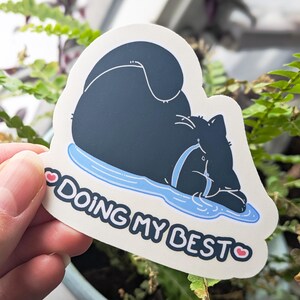 May include: A die-cut sticker showing a black cat resting on a blue puddle. The sticker reads "DOING MY BEST" in white text, with a pink heart on each side. The cat is wearing a blue collar.