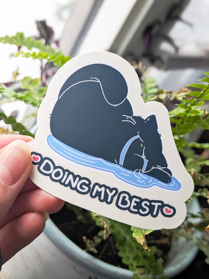 May include: A die-cut sticker showing a black cat resting on a blue puddle. The sticker reads "DOING MY BEST" in white text, with a pink heart on each side. The cat is wearing a blue collar.